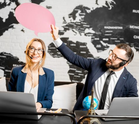 Couple of travel managers dreaming with colorful bubbles above the head sitting at the agency office with world map on the background